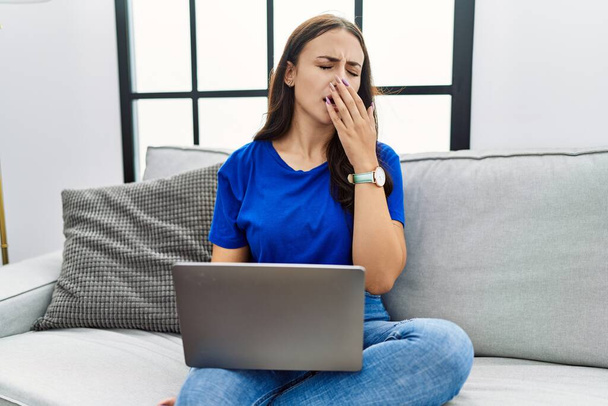 Νεαρή μελαχρινή γυναίκα που χρησιμοποιεί φορητό υπολογιστή στο σπίτι βαριέται χασμουρητό κουρασμένος καλύπτει το στόμα με το χέρι. ανησυχία και υπνηλία.  - Φωτογραφία, εικόνα