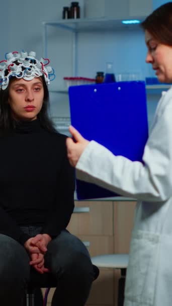 Verticale video: Neuroscience arts die klembord toont behandeling tegen hersenziekte aan patiënt met eeg headset. Vrouw in neurologisch wetenschappelijk laboratorium die het zenuwstelsel behandelt - Video