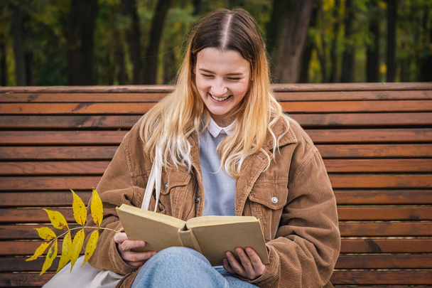 Eine attraktive junge Frau sitzt auf einer Holzbank im herbstlichen Park und liest ein Buch. - Foto, Bild