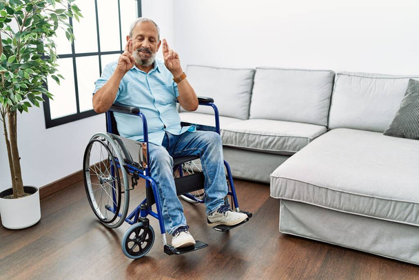 Pohledný starší muž sedící na vozíčku v obývacím pokoji a ukazující prstem, zkříženě se usmíval s nadějí a zavřenýma očima. štěstí a pověrčivý koncept.  - Fotografie, Obrázek