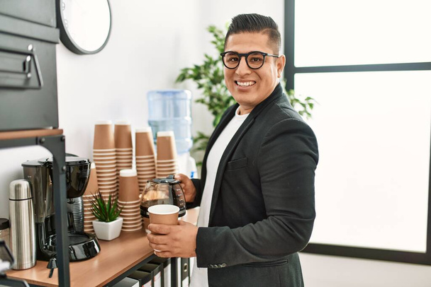 Νεαρός Λατίνος επιχειρηματίας χαμογελώντας χαρούμενος πίνοντας καφέ στο γραφείο. - Φωτογραφία, εικόνα