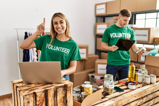 Молодая женщина и мужчина в футболке добровольца на пожертвования стенд удивлен идеей или вопрос указывая пальцем со счастливым лицом, номер один  - Фото, изображение