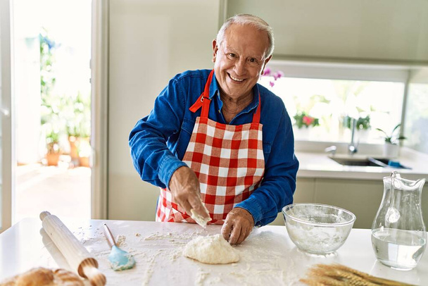 Homem sênior sorrindo confiante derramando farinha na massa na cozinha - Foto, Imagem