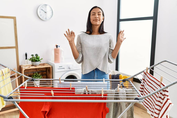 Young hispanic woman putting fresh laundry on clothesline relax and smiling with eyes closed doing meditation gesture with fingers. yoga concept.  - Photo, Image