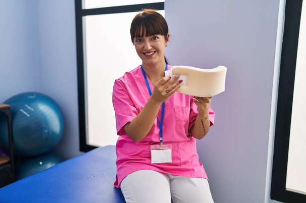 Νεαρή όμορφη ισπανίδα ψυχοθεραπεύτρια χαμογελά με αυτοπεποίθηση κρατώντας τον αυχενικό λαιμό στην κλινική αποτοξίνωσης - Φωτογραφία, εικόνα