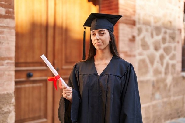Young hispanic woman wearing graduated uniform holding diploma at university - Photo, image