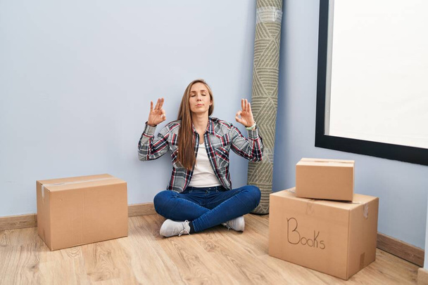 Mujer rubia joven sentada en el suelo moviéndose a un nuevo hogar relajarse y sonreír con los ojos cerrados haciendo gesto de meditación con los dedos. concepto de yoga.  - Foto, Imagen
