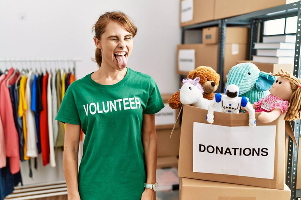Beautiful caucasian woman wearing volunteer t shirt at donations stand sticking tongue out happy with funny expression. emotion concept.  - Photo, Image