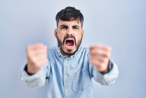 Joven hombre hispano con barba de pie sobre fondo azul enojado y loco levantando puños frustrado y furioso mientras gritaba con ira. rabia y concepto agresivo.  - Foto, imagen