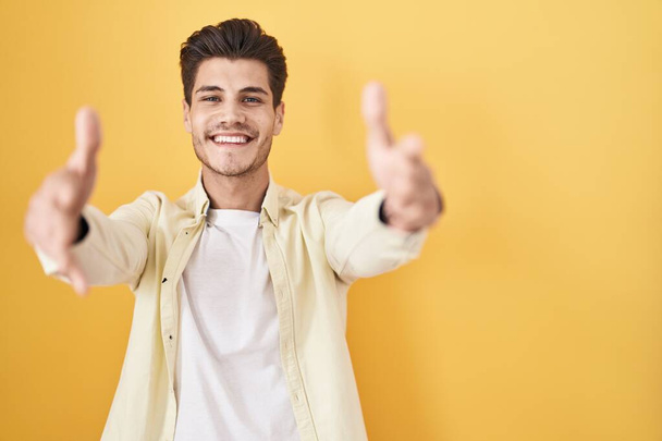 Jeune homme hispanique debout sur fond jaune regardant la caméra souriant à bras ouverts pour un câlin. expression joyeuse embrassant le bonheur.  - Photo, image
