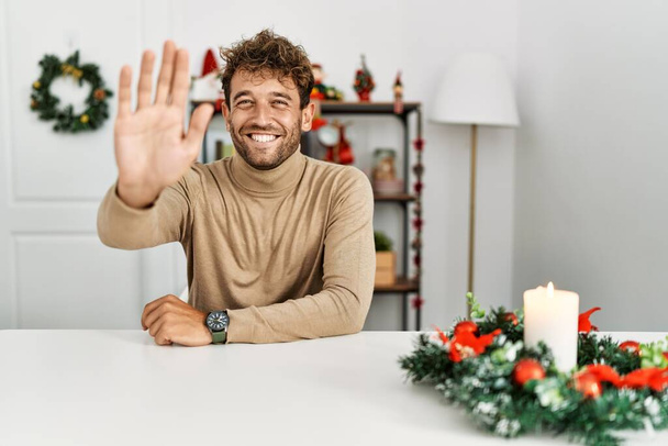 ひげを生やした若いハンサムな男はクリスマスの装飾によってテーブルの上に座って挨拶をしながら幸せと笑顔、優しい歓迎ジェスチャー  - 写真・画像