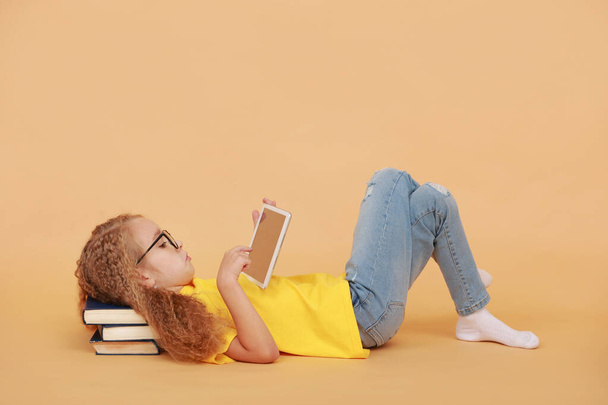 Entzückendes Schulmädchen mit lockigem Haar in Brille hält ein digitales Tablet in der Hand und liegt auf Papierbüchern isoliert auf gelbem Studiohintergrund. Kinder entscheiden sich für moderne Technologie-Bildung statt für traditionelle. - Foto, Bild