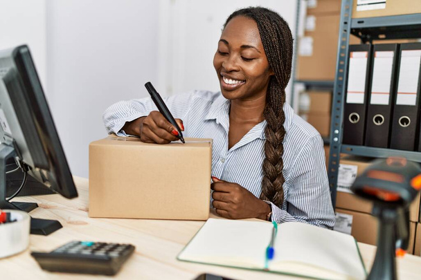 Afrika kökenli Amerikalı kadın ecommerce işçisi ofiste paket üzerinde bilgisayar kullanıyor. - Fotoğraf, Görsel