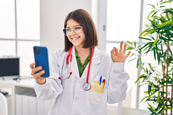 Молодая латиноамериканка доктор женщина, работающая на сайте назначения делает хорошо знак с пальцами, улыбаясь дружелюбный жест отличный символ  - Фото, изображение