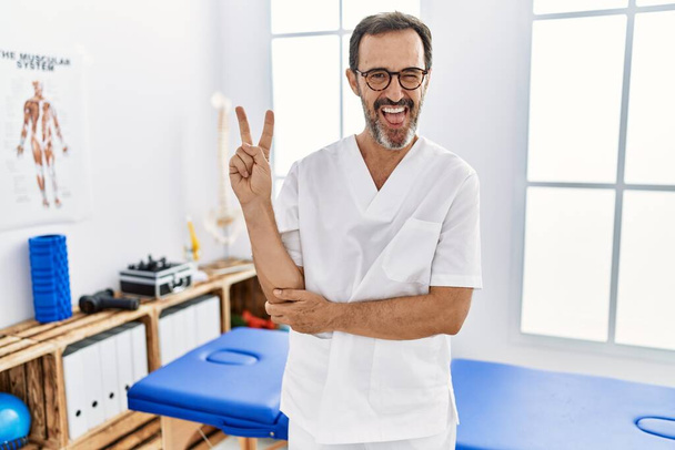 Middle age man with beard working at pain recovery clinic smiling with happy face winking at the camera doing victory sign. number two.  - Zdjęcie, obraz