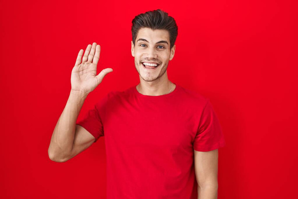 Mladý hispánec stojící nad červeným pozadím a vzdávající se pozdravu šťastný a usmívající se, přátelské gesto  - Fotografie, Obrázek