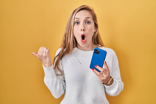 Giovane donna bionda utilizzando smartphone digitando messaggio sorpreso puntando con il dito della mano di lato, bocca aperta espressione stupita.  - Foto, immagini