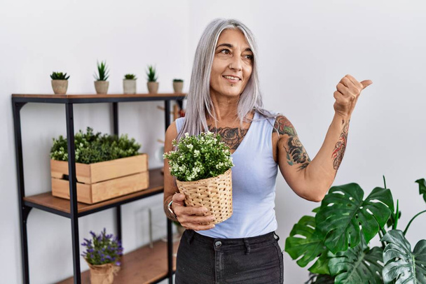 Mujer de pelo gris de mediana edad sosteniendo maceta verde en casa sonriendo con la cara feliz mirando y señalando a un lado con el pulgar hacia arriba.  - Foto, imagen