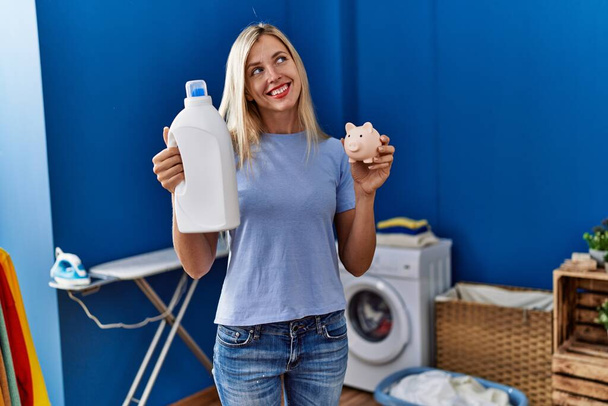 Beautiful woman doing laundry holding detergent bottle and piggy bank smiling looking to the side and staring away thinking.  - Photo, Image