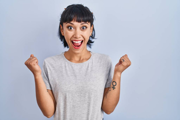 Young hispanic woman wearing casual t shirt over blue background celebrating surprised and amazed for success with arms raised and open eyes. winner concept.  - Photo, Image