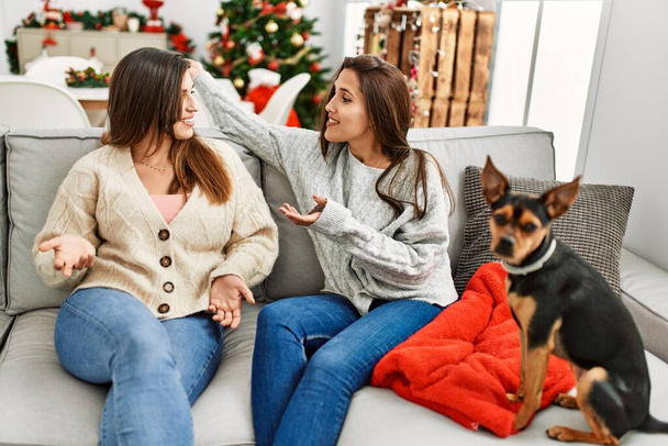 Two women having conversation sitting with dog by christmas tree at home - Photo, Image