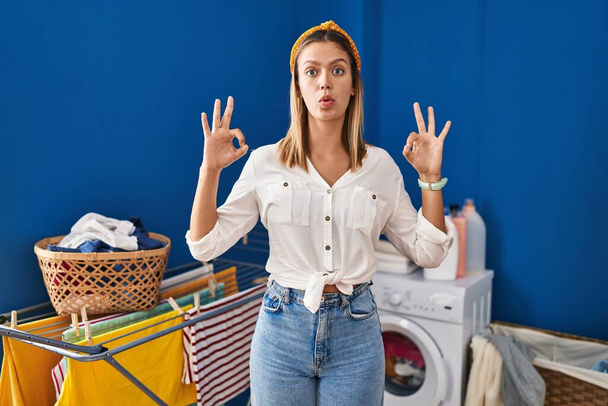 Giovane donna bionda in lavanderia guardando sorpreso e scioccato facendo ok simbolo di approvazione con le dita. espressione folle  - Foto, immagini