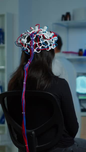 Vidéo verticale : Vue arrière de la patiente portant un casque eeg performant assis sur la chaise dans un laboratoire de recherche neurologique pendant que le chercheur médical l'ajuste, examinant le système nerveux tapant sur - Séquence, vidéo