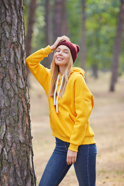 Attractive blue eyed blonde woman walk on the park. Girl wear yellow hoodie and burgundy hat, look happy and smiles. Portrait of a joyful young woman enjoying in autumn park. - Photo, Image