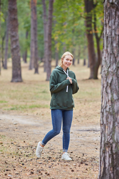 Cheerful blue-eyed blonde girl in green hoodie, smiles against park background. Attractive blonde in stylish clothes posing outdoor. Relax in nature. - Фото, зображення