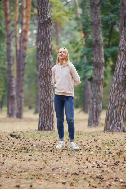 ベージュ色のパーカーの青い目のブロンドは、松林の中を歩く。秋の公園で楽しむ楽しい若い女性の肖像画。自然の中でリラックス. - 写真・画像