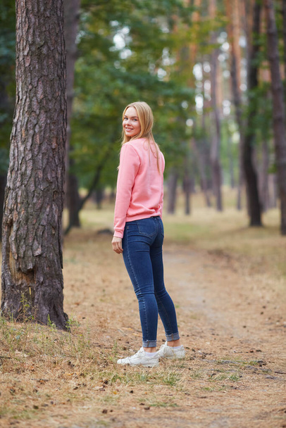 Блакитноока блондинка в рожевому светрі гуляє в сосновому лісі. Портрет щасливої молодої жінки, яка насолоджується осіннім парком. Відпочинок в природі
. - Фото, зображення