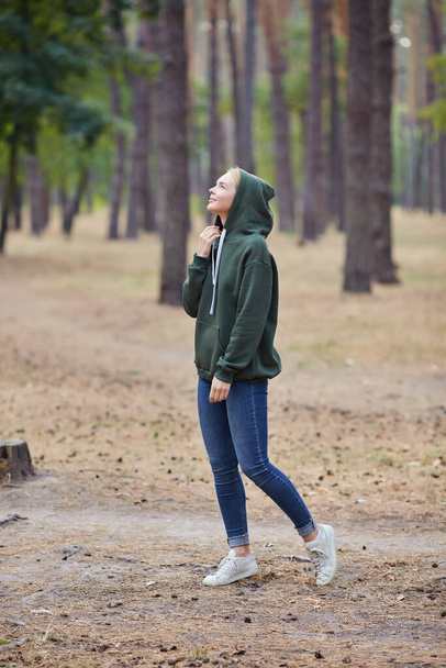 Блакитноока блондинка в зеленому светрі гуляє в сосновому лісі. Портрет щасливої молодої жінки, яка насолоджується осіннім парком. Відпочинок в природі
. - Фото, зображення
