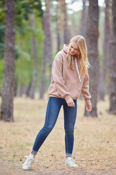 Mooie jonge blonde vrouw in beige hoodie wandelingen in het park. Aantrekkelijke vrouw die actief tijd doorbrengt in het park. Portret van een vrolijke jonge vrouw die geniet in het herfstpark. Ontspan in de natuur. - Foto, afbeelding