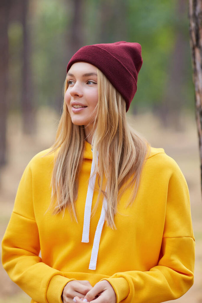 松の森の中にある黄色いパーカーとバーガンディの帽子の青い目のブロンド。秋の公園で楽しむ楽しい若い女性の肖像画。自然の中でリラックス. - 写真・画像
