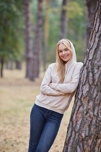 Attractive blue eyed blonde woman walk on the city park. Girl wear beige hoodie, pink bag and look happy and smiles. Portrait of a joyful young woman enjoying in autumn park. Relax in nature. - Фото, изображение