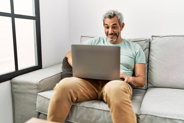 Grauhaariger Mann mittleren Alters sitzt mit Laptop auf Sofa zu Hause - Foto, Bild