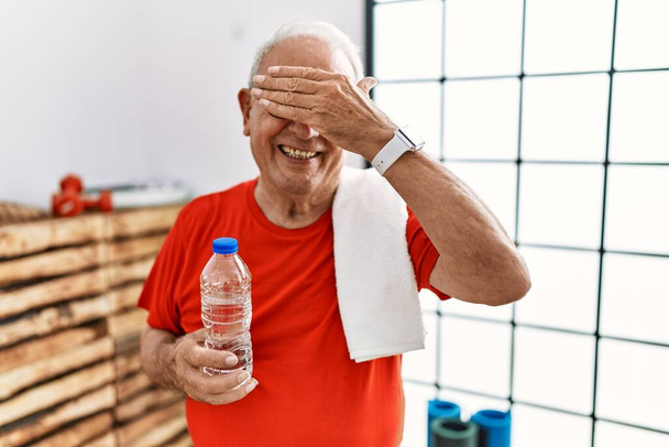 Старший мужчина в спортивной одежде и полотенце в тренажерном зале улыбается и смеется, закрывая глаза рукой. слепая концепция.  - Фото, изображение
