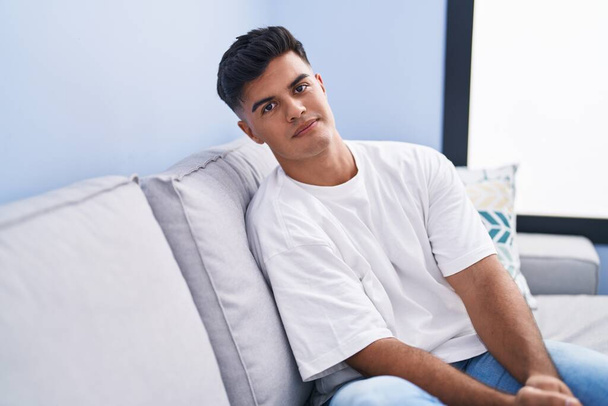 Young hispanic man sitting on sofa with relaxed expression at home - Foto, afbeelding