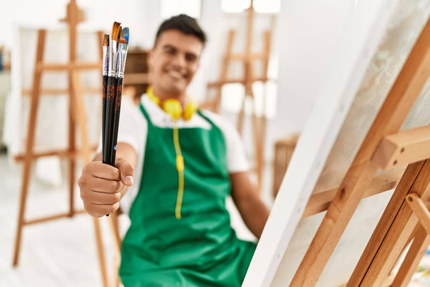Young hispanic man smiling confident holding paintbrushes at art studio - Zdjęcie, obraz