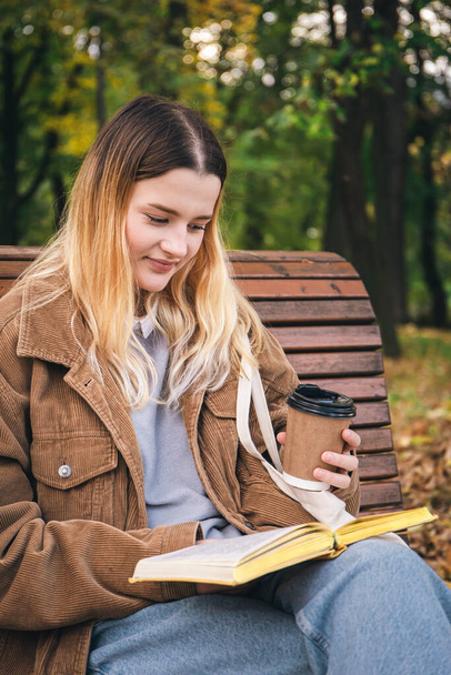 Atrakcyjna młoda kobieta siedzi na drewnianej ławce w jesiennym parku, pije kawę i czyta książkę.. - Zdjęcie, obraz
