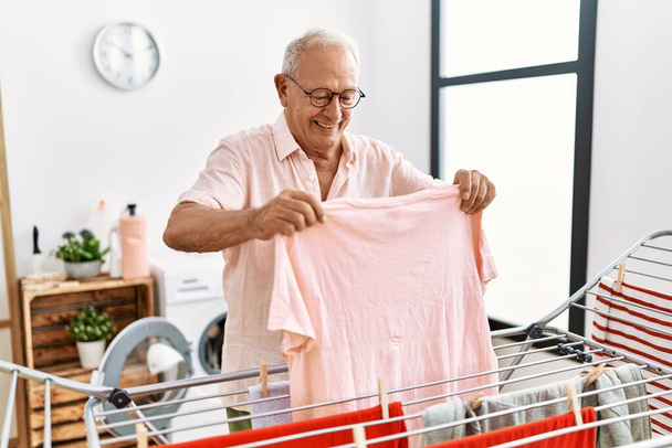 Uomo anziano sorridente fiducioso appeso vestiti su clothesline in lavanderia - Foto, immagini
