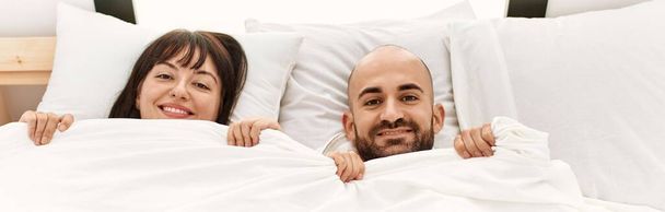 Young hispanic couple covering face with bedsheet lying on the bed at bedroom. - Фото, изображение