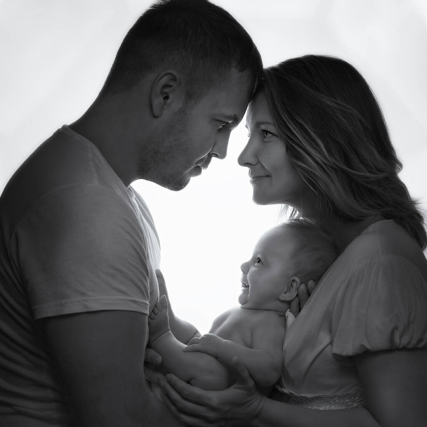 Ασπρόμαυρη φωτογραφία ευτυχισμένης οικογένειας με τον γιο του βρέφους δίπλα στο παράθυρο. Χαμογελώντας νεογέννητο μωρό στην αγκαλιά της μαμάς και του μπαμπά. Έννοια της αγάπης, προστασία των παιδιών. - Φωτογραφία, εικόνα