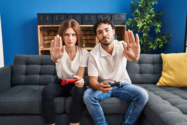 Junges Paar spielt Videospiel mit Controller zu Hause mit offener Hand macht Stoppschild mit ernstem und selbstbewusstem Gesichtsausdruck, Verteidigungsgeste  - Foto, Bild
