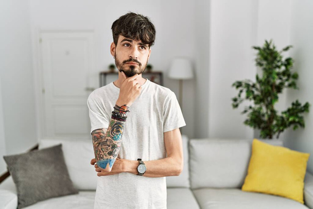 Ein spanischer Mann mit Bart zu Hause im Wohnzimmer, die Hand am Kinn, über Fragen nachdenkend, nachdenklich. Lächeln mit nachdenklichem Gesicht. Zweifelhaftes Konzept.  - Foto, Bild