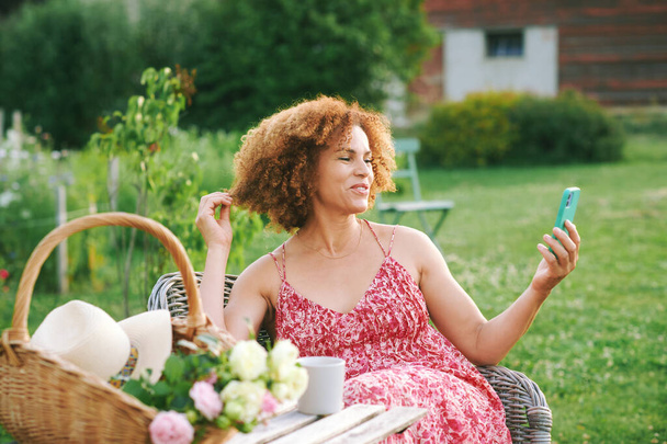 Outdoor portrait of beautiful mature woman resting in summer garden, sitting in cosy chair, having video call with her family or friend, basket with freshly cut rosses standing on the table - Фото, изображение