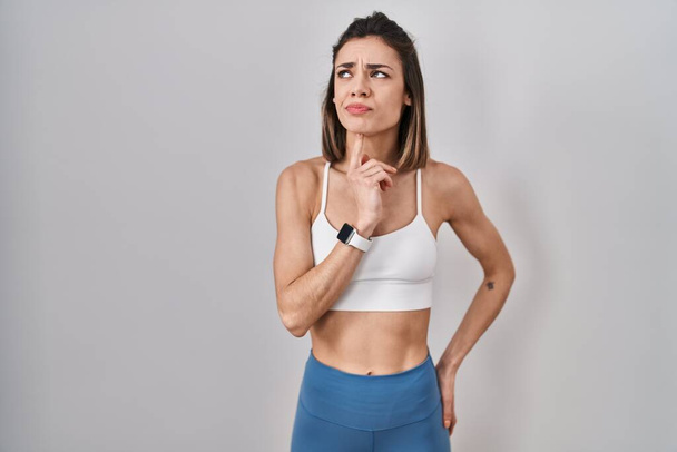 Латиноамериканка в спортивной одежде на изолированном фоне думает о сомнениях с пальцем на подбородке и смотрит вверх, удивляясь:  - Фото, изображение