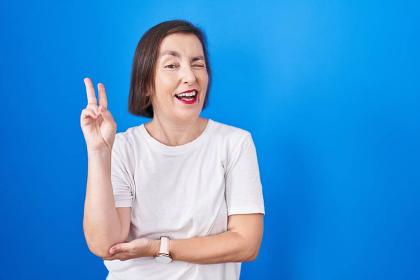 Middelbare leeftijd Spaanse vrouw die over een blauwe achtergrond staat te glimlachen met een vrolijk gezicht knipogen naar de camera doen overwinning teken met de vingers. nummer twee.  - Foto, afbeelding