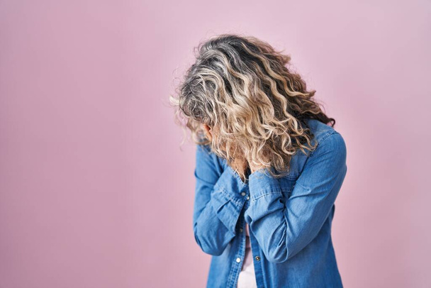 Μεσήλικας γυναίκα στέκεται πάνω από ροζ φόντο με θλιβερή έκφραση που καλύπτει το πρόσωπο με τα χέρια, ενώ κλαίει. έννοια της κατάθλιψης.  - Φωτογραφία, εικόνα