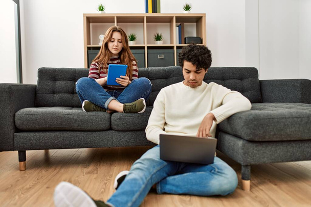 Młoda para z poważnym wyrazem twarzy za pomocą laptopa i touchpad siedzi na kanapie w domu. - Zdjęcie, obraz